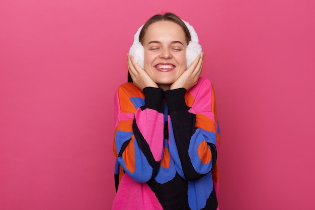 Image of smiling satisfied delighted Caucasian woman wearing warm sweater and fur earmuffs standing isolated over pink background smiling happily closed her eyes enjoying moment