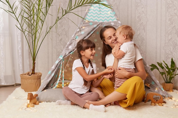 Immagine di bambine sorridenti soddisfatte e sua madre che giocano a casa a wigwam ed essere felici di trascorrere del tempo insieme abbracciando i suoi figli essendo di buon umore