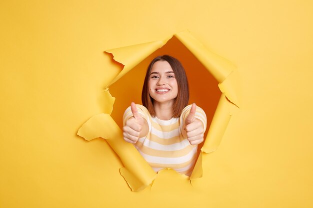 Image of smiling satisfied young woman wearing striped shirt showing thumbs up positive feedback recommend service looking through hole in yellow paper