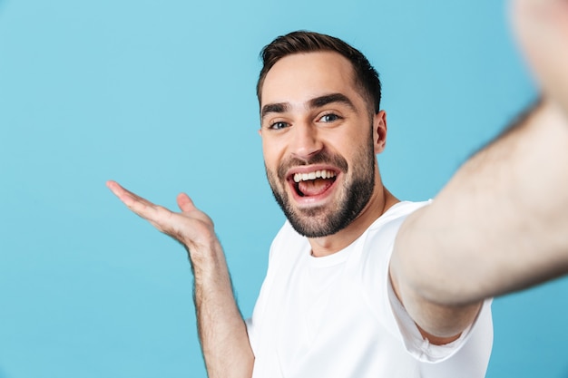 Image of smiling young bearded man posing isolated over blue wall  take a selfie by camera showing copyspace.