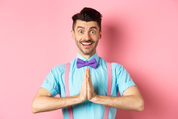 Image of smiling young man asking please, looking grateful at
camera, guy in bow-tie say thank you, standing over pink.