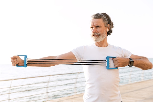 Image of strong mature man in sportswear using earpod while doing exercise with expander near seaside in morning