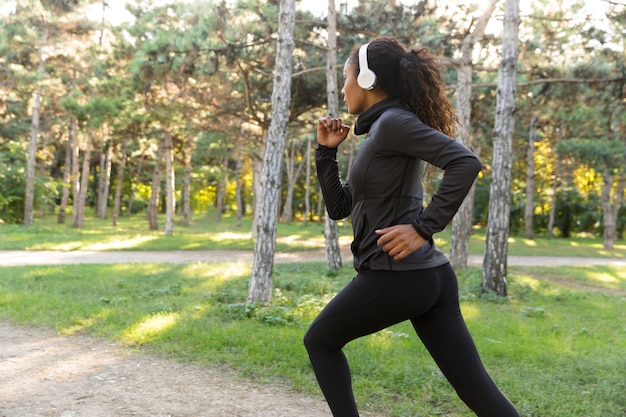 Image of strong woman 20s wearing black tracksuit and headphones working out, while running through green park