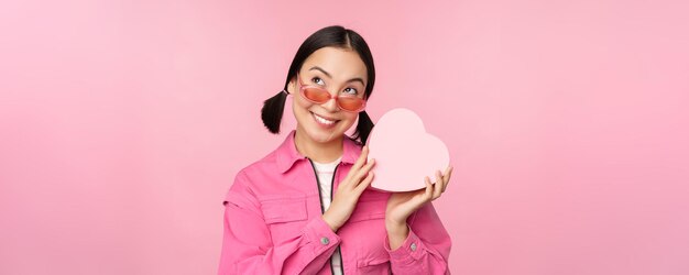 Image of stylish asian girlfriend in sunglasses guessing whats inside gift box heart-shaped present standing over pink background person