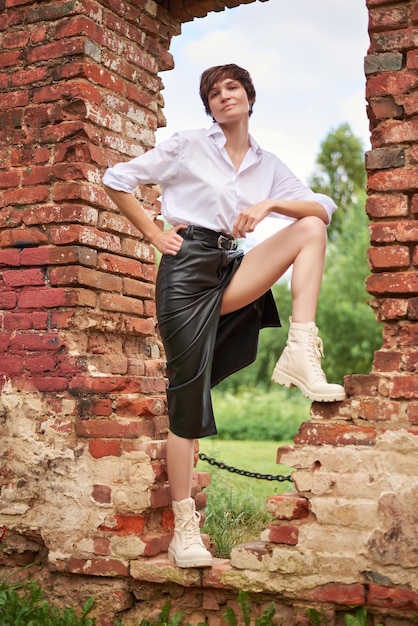 Image of a stylish beautiful woman in a white shirt and leather skirt in a park against the background of a destroyed building The concept of style and fashion