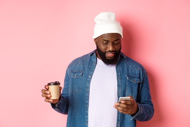 Image of stylish Black man hipster drinking takeaway coffee, reading message on phone, standing over pink background.