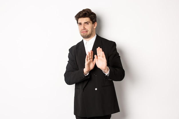 Image of stylish handsome man in black suit, cringe from something embarrassing, declining offer and showing stop gesture, standing against white background