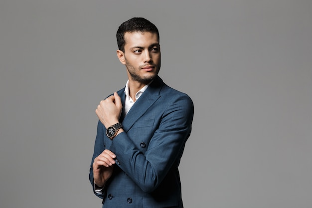 灰色の壁に隔離された、脇を向いて腕時計を示すフォーマルなスーツを着た30代の成功したアラビアのビジネスマンの画像