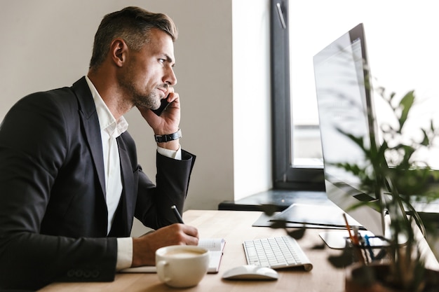 Image of successful businessman 30s wearing suit talking on cell phone while working on computer in office