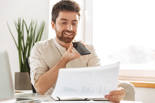 Immagine di imprenditore di successo 30s indossa una camicia bianca parlando al telefono cellulare e tenendo un documento cartaceo, mentre si lavora in ufficio