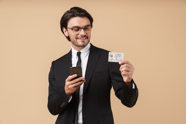 Image of successful businessman in formal suit holding cellphone and credit card isolated over beige wall