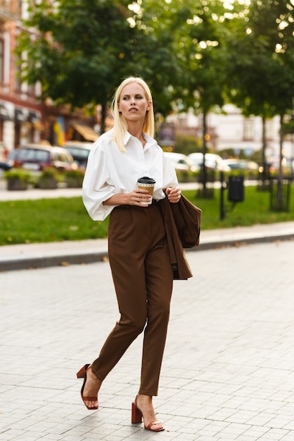 Photo image of successful elegant woman wearing white shirt drinking coffee takeaway while walking at summer boulevard