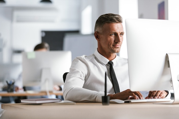 Image of successful employee man 30s wearing white shirt and tie sitting at desk in office, and working at computer