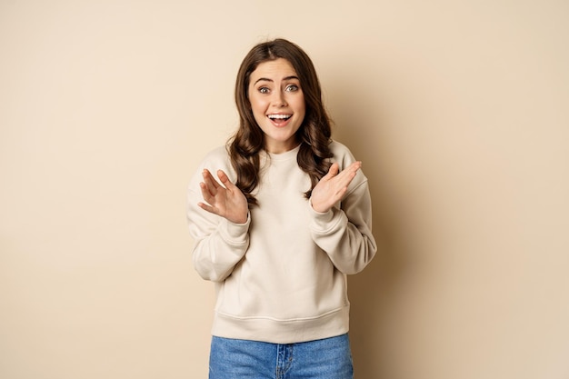 Image of surprised amazed brunette girl gasping looking fascinated at camera standing over beige bac...