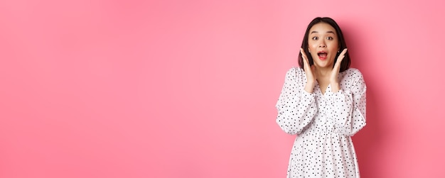 Image of surprised korean girl in dress female model staring at camera and gasping amazd standing ov