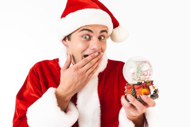 Image of surprised man 30s in santa claus costume and red hat holding christmas ball