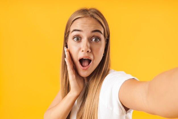 Photo image of surprised woman wearing basic clothes screaming in wonder while taking selfie photo isolated over yellow wall