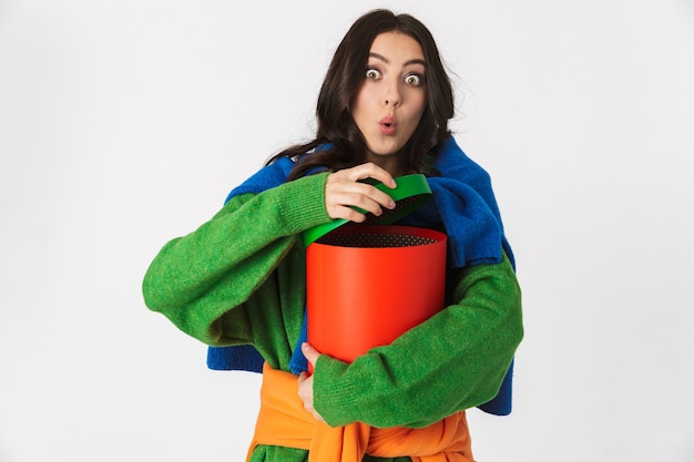 Image of surprised woman with dark hair in colorful clothes holding big gift box while standing, isolated on white