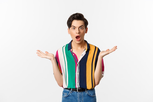 Image of surprised young male model staring at camera, spread hands sideways in complete disbelief, standing over white background.