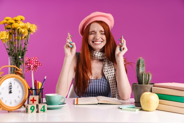 Image of tense teenage girl keeping fingers crossed while studying with exercise books isolated over violet wall