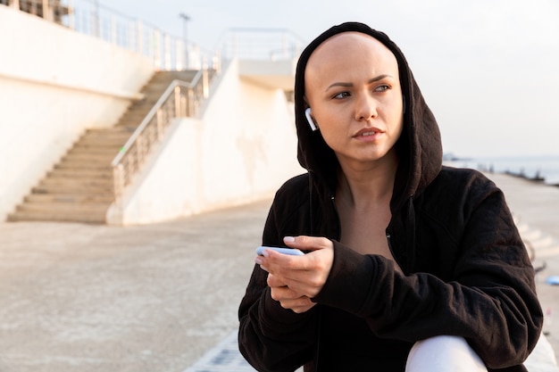 Image of thinking bald woman in black hood using cellphone and earphone while sitting on boardwalk in sunny morning