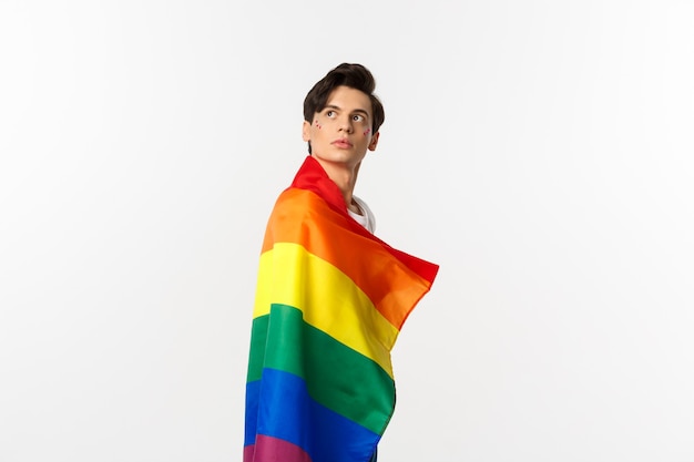 Immagine del giovane gay premuroso che indossa la bandiera lgbtq, gira la testa nell'angolo in alto a sinistra, fissando il logo, in piedi sopra il bianco.
