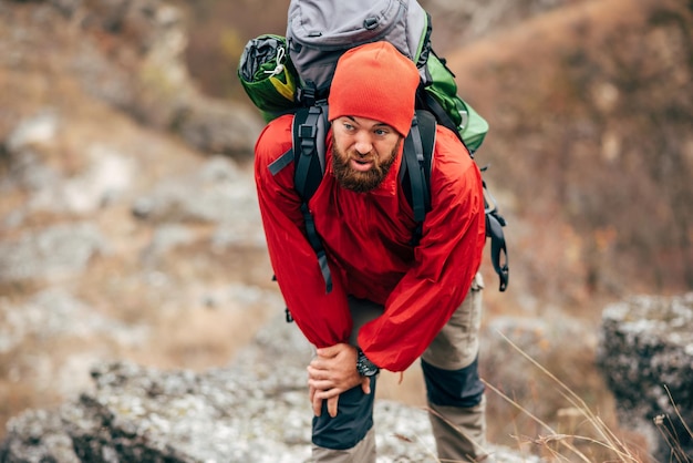 Image of tired hiker man hiking in mountains dressed in red clothes exploring new places Traveler bearded male trekking and mountaineering during his journey Travel people sport lifestyle concept