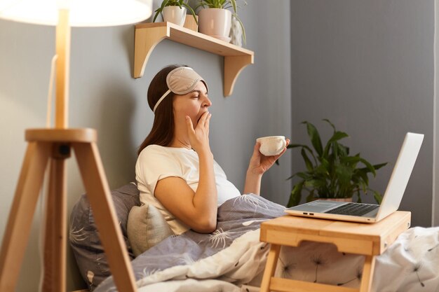 Image of tired sleepless caucasian woman wearing sleep mask sitting with laptop in bed at home holding cup of coffee or tea looking at notebook screen and yawning covering mouth with hand