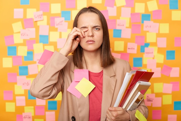 Image of tired stressed young adult woman holding papers posing\
against yellow wall with colorful stickers rubbing her eye feels\
pain looking at camera with pout lips