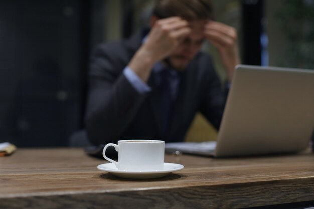 Image of tired young business man in suit keeping face in hands while sitting in the office.