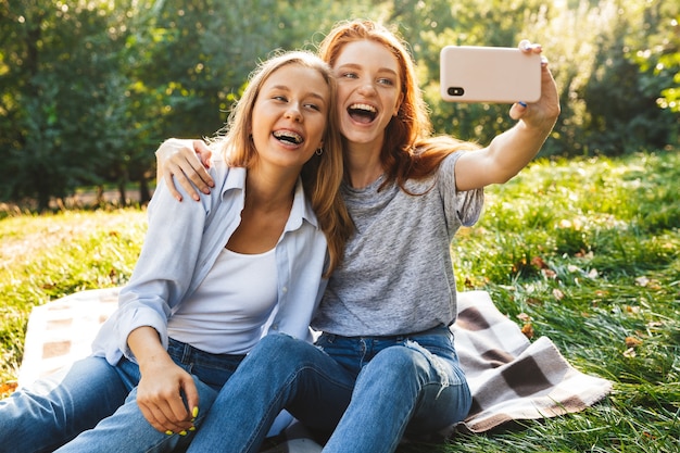 Image of two joyful women wearing casual clothes laughing, while sitting on grass and taking selfie on mobile phone, while resting in summer park