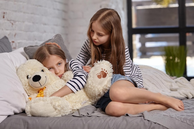 The image of two little sisters sitting on the bed in the room