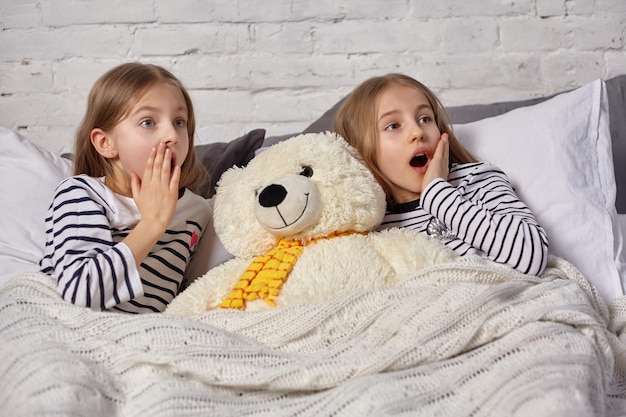 The image of two little sisters waking up in the morning. Big white plush toy bear laying between them. Something scared them