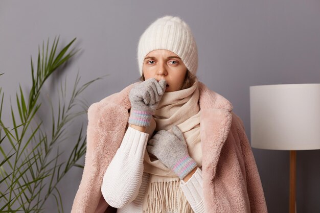 Image of unhealthy sick Caucasian woman wearing cap and coat posing in living room and coughing suffering flu and sore throat symptoms being in frozen room