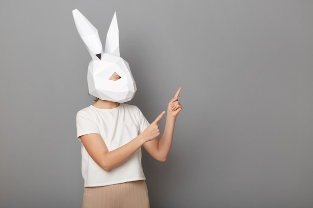 Image of unknown woman wearing white t shirt and paper rabbit mask standing isolated over gray background pointing aside at advertisement area copy space for your promotional text