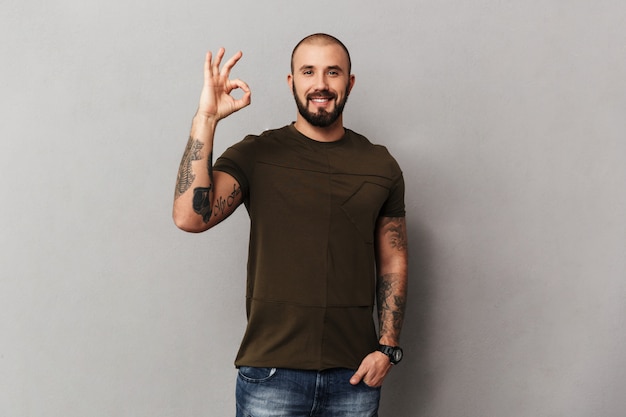 Image of unshaved muscular guy with tattoos on hands posing  and showing OK sign, isolated over gray wall