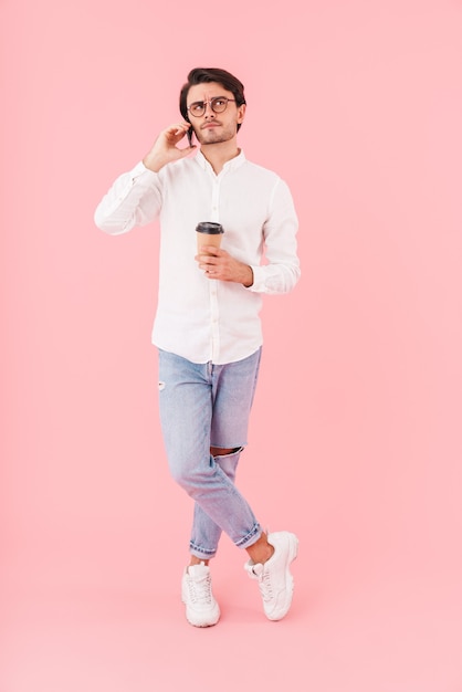 Image of unshaven perplexed man wearing eyeglasses drinking coffee takeaway and talking on cellphone isolated