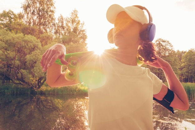 Image of a woman in a cap and headphones with a skateboard. Sunset in the park. Vacation concept. Mixed media