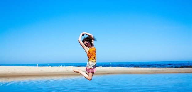 白い砂丘と青い空を背景に明るい青い湖でジャンプする暗い巻き毛の女性の画像