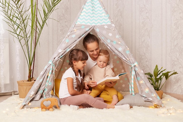 Image of woman with her little daughters reading book in hovel at home Caucasian female sitting on floor in wigwam read fairy tale to her kids spending happy time together