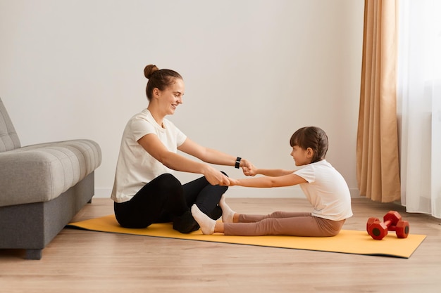 Image of woman with little girl wearing sportswear doing sport exercises at home sitting on floor holding hands together and practicing yoga at home doing stretching exercise