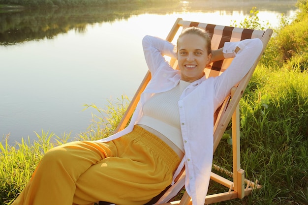 Image of young adult woman with toothy smile sitting in the folding chair at the picturesque bank of the river looking at camera and keeps hands behind head resting outdoors