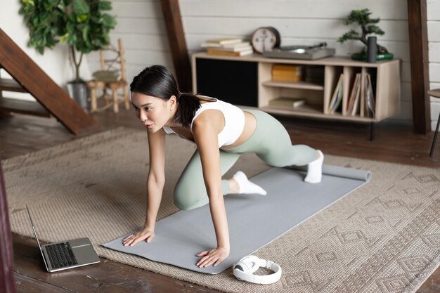 Photo image of young asian girl doing workout at home following fitness instructor online on laptop doing ...