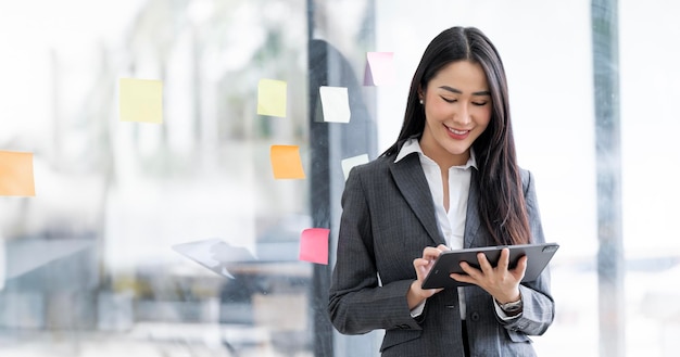 Изображение молодой азиатки, улыбающейся и держащей в руках цифровой планшет, стоящий в офисе