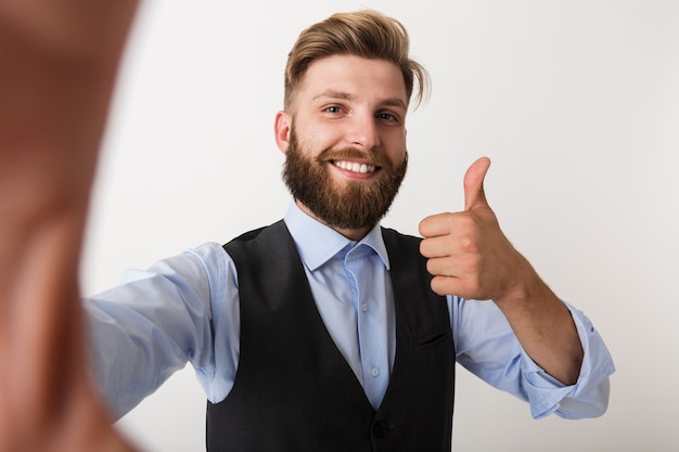 Photo image of a young bearded man standing isolated over white wall make selfie showing thumbs up.