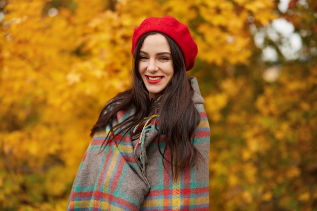 Image of young beautiful brunette woman wrapped in checkered blanket or plaid walks outdoors through autumn park surrounded