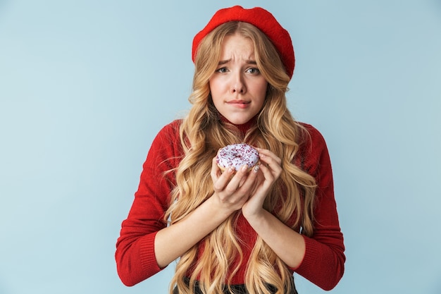 Image of young blond woman 20s wearing red beret eating donut isolated 