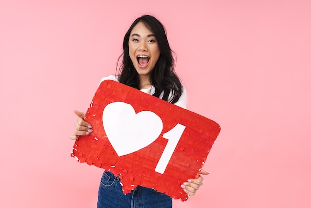 Image of young brunette asian woman wearing eyeglasses holding heart like symbol on placard isolated on pink