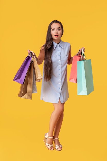 Image of a young brunette lady in white and blue summer striped dress posing with shopping bags and looking at camera over yellow background.