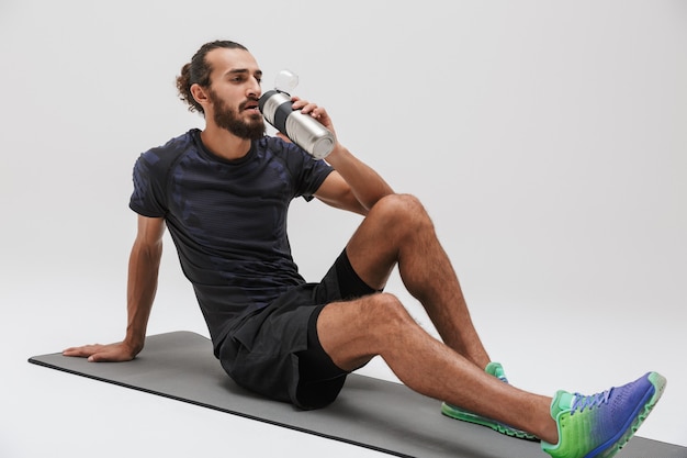 Image of young brunette sportsman in tracksuit drinking water while doing workout on yoga mat